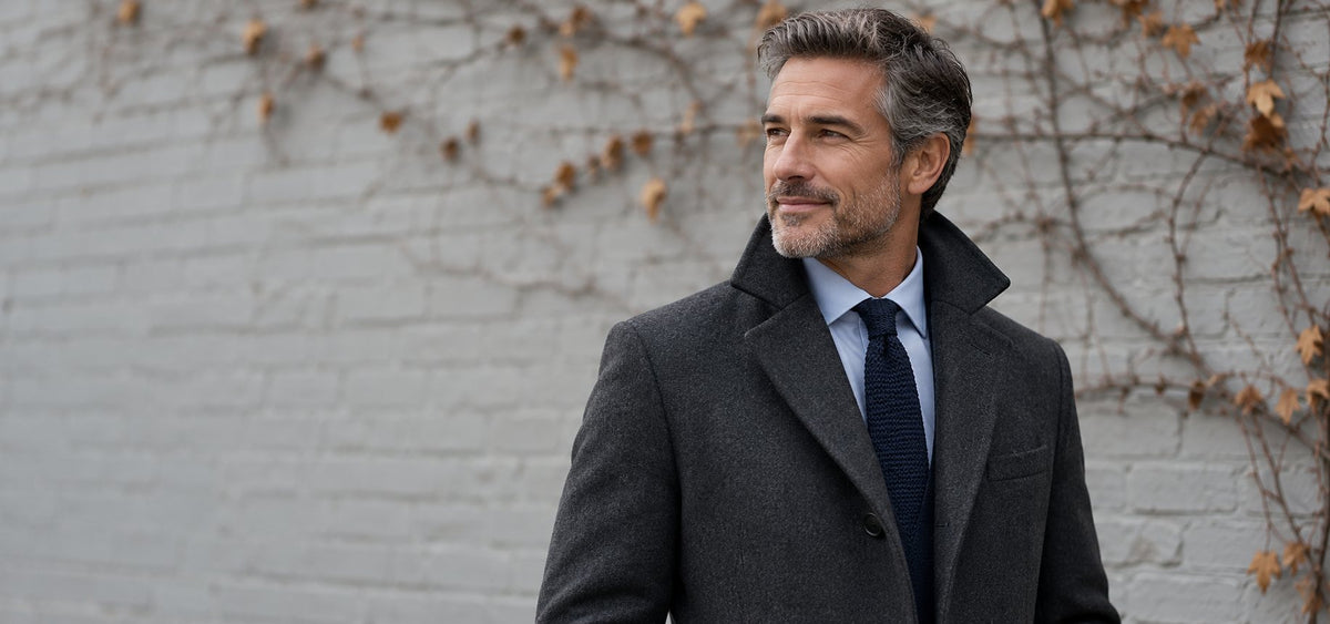 Man in a charcoal wool overcoat and dress shirt against a painted brick wall – ZOCOIFY Fall '26